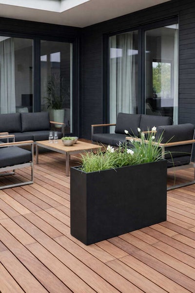 Moderne Holzterrasse mit großem schwarzem rechteckigem Pflanzkübel, dunkelgrauen Loungemöbeln und einem Holztisch in einer hellen Außenanlage.
