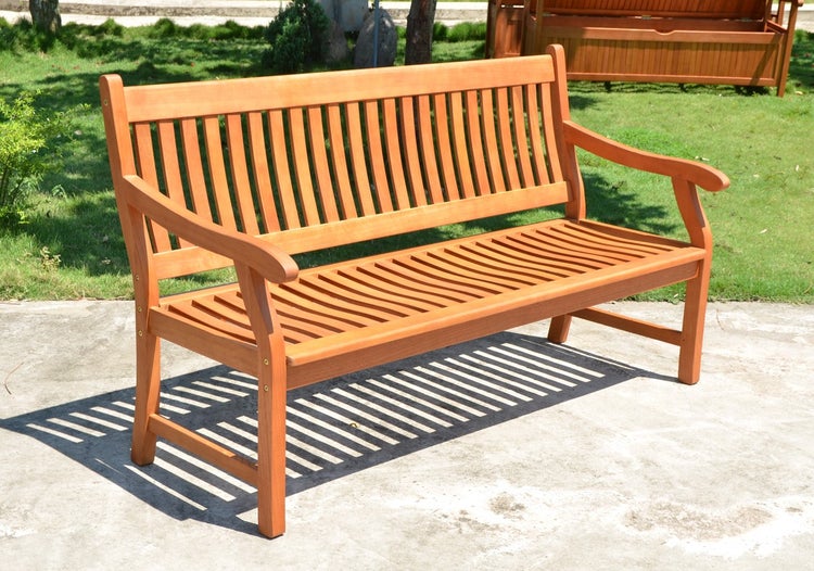 Gartenbank aus Holz mit Armlehnen und Rückenlehne mit vertikalen Latten auf einer hellen Terrasse im Garten.