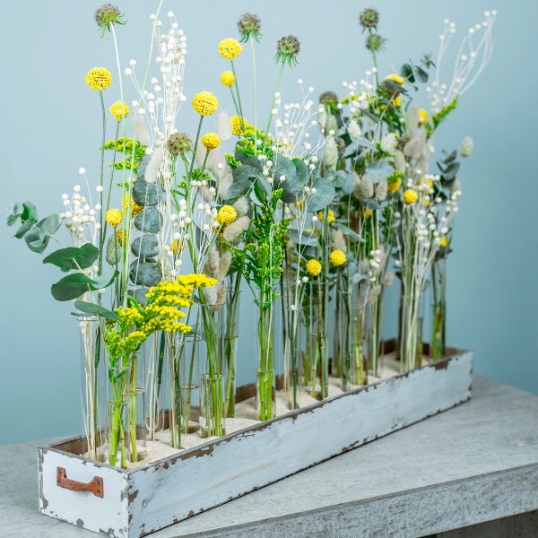 Gesteck aus Trockenblumen mit gelben Blüten und Eukalyptus in Glasröhrchen auf einem rustikalen weißen Holztablett.