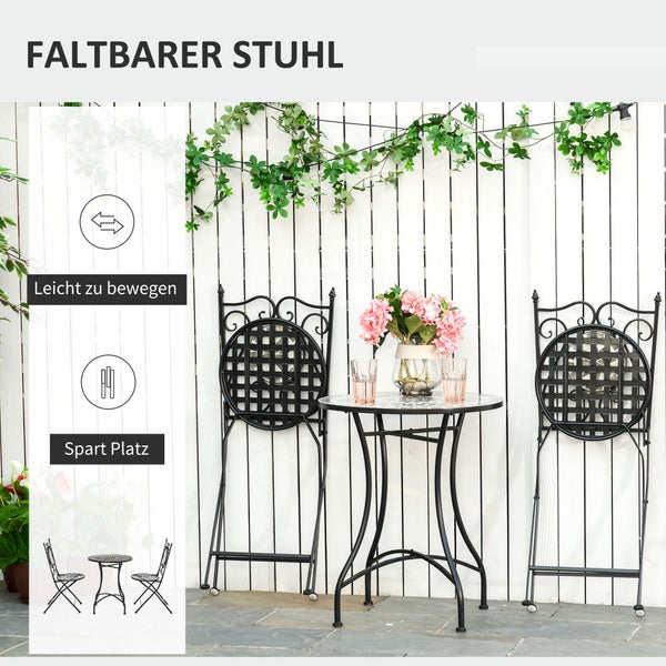 Zusammengeklappte Gartenstühle aus schwarzem Metall mit Ornamenten und runder Bistrotisch vor weißem Zaun. Platzsparend und leicht beweglich.