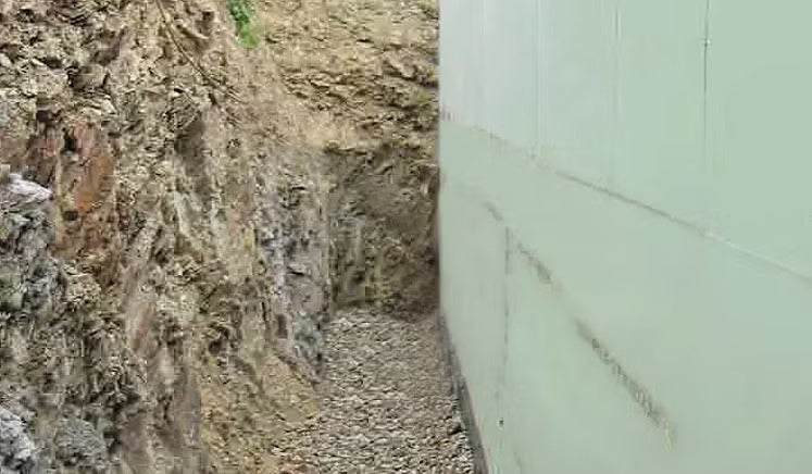 Baugrube mit einer gedämmten Hauswand und einer Kiesschicht im Graben neben einer Erdwand.
