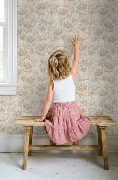 Ein kleines Kind sitzt auf einer Holzbank vor einer Wand mit floraler Mustertapete in einem hellen, freundlich eingerichteten Raum.