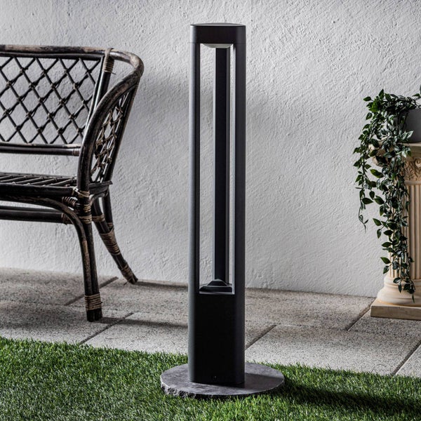 Moderne Wegeleuchte auf Steinsockel im Garten neben Rattansessel und Pflanze vor einer strukturierten Wand.