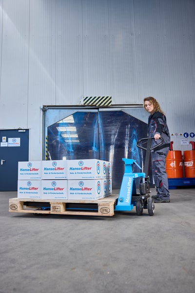 Eine Frau in Arbeitskleidung bedient einen HanseLifter Hubwagen mit einer Europalette voller Kartons in einer hellen Lagerhalle.