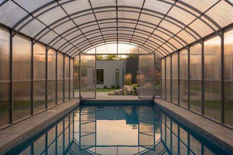Überdachter Außenpool mit gewölbter Polycarbonat Poolüberdachung vor einer modernen Wohnhausfassade.