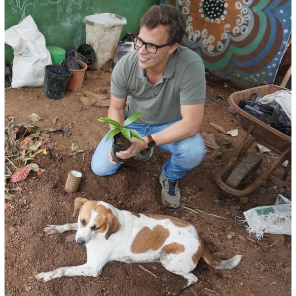 Mann hockt mit Baumsetzling und Hund im Garten.