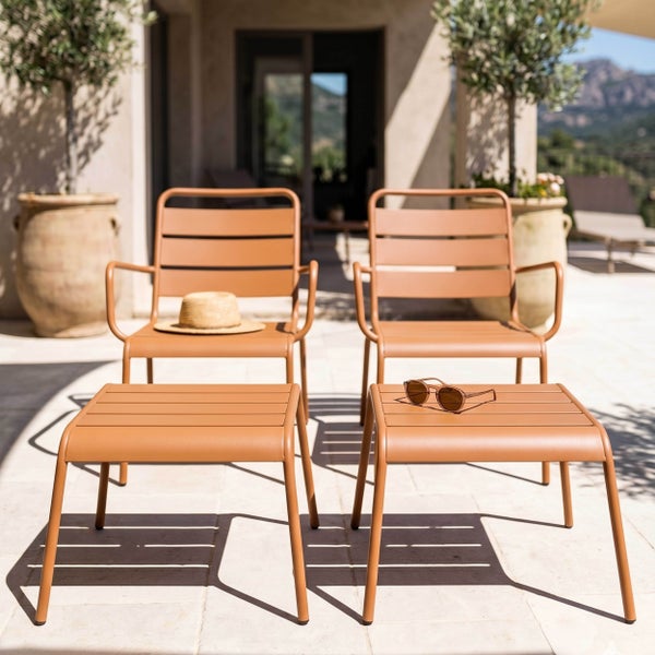 Zwei moderne Metall-Gartenstühle und zwei passende Hocker in Terracotta auf einer hellen Terrasse mit Olivenbäumen in Töpfen.