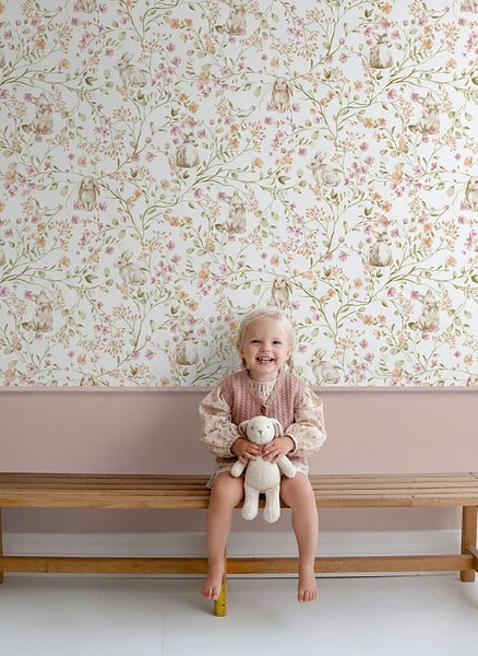 Vliestapete mit Hasen- und Blumenmuster im Aquarell-Stil an einer Kinderzimmerwand über einer rosa Wandfläche und einer Holzbank.
