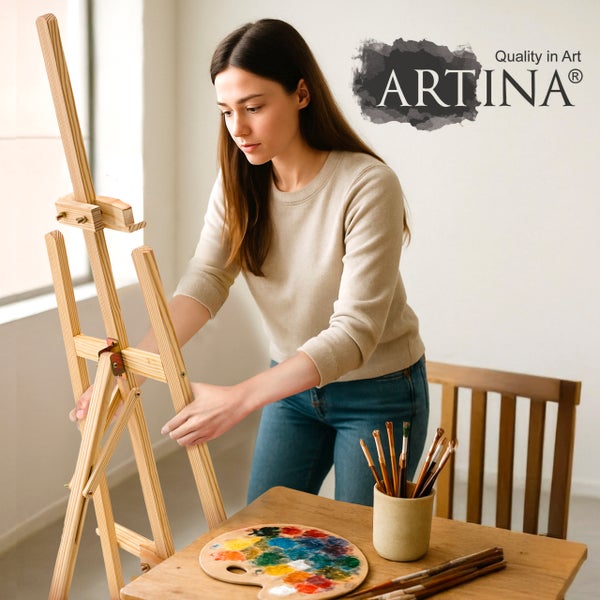 Frau stellt hölzerne Artina Dreibeinstaffelei in hellem Raum auf, mit Palette und Pinseln auf einem Tisch. Artina Logo.