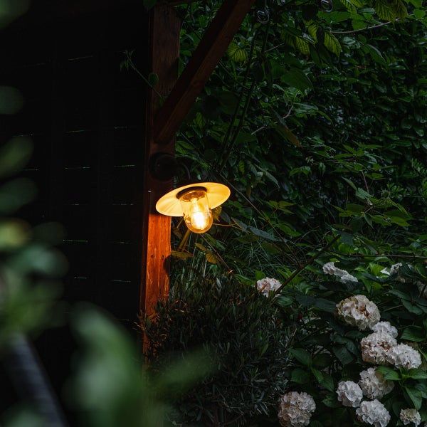 Außenwandleuchte mit warmem Licht an einem Holzpfosten in einem Garten mit grünem Laub und weißen Hortensien.