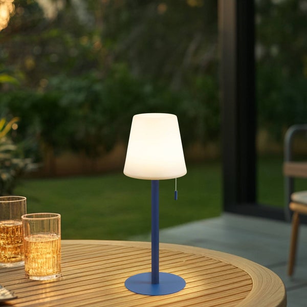 Moderne kabellose LED-Außentischleuchte mit blauem Fuß und weißem Schirm auf einem hölzernen Gartentisch.
