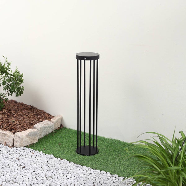 Moderne schwarze Wegeleuchte in Säulenform mit vertikalen Streben auf grünem Kunstrasen neben einem Kieselweg.