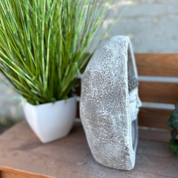 Dekorative Buddha-Kopf-Skulptur in Steinoptik, von der Seite fotografiert, neben einer künstlichen Graspflanze im Topf auf einer Holzbank.