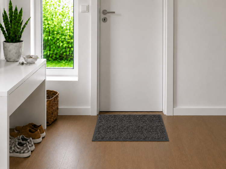 Dunkelgraue Fußmatte vor einer weißen Innentür in einem hellen Flur mit Holzboden, weißer Konsole und einer Zimmerpflanze.