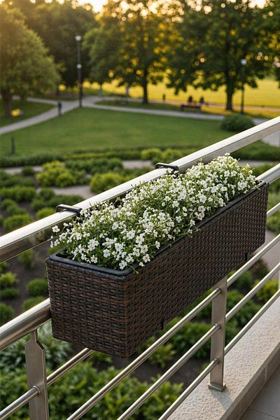 Rechteckiger Balkonkasten in brauner Rattan-Optik mit weißen Blumen an einem Metallgeländer. Im Hintergrund eine Parkanlage.