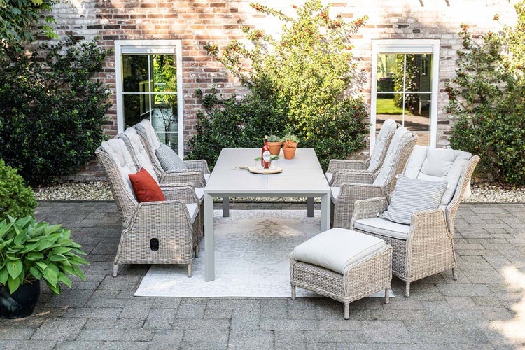 Gartenmöbel-Set aus Polyrattan-Geflecht mit Esstisch, vier Sesseln inklusive Polstern und einem Hocker auf einer gepflasterten Terrasse.