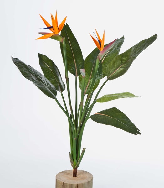 Künstliche Strelitzia mit zwei orangefarbenen Blüten und großen grünen Blättern auf einem Holzsockel.