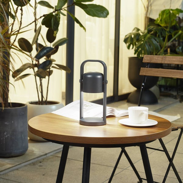 Tragbare LED-Tischleuchte auf einem runden Holztisch neben einer Kaffeetasse und einer Zeitschrift in einer hellen Umgebung mit Zimmerpflanzen.