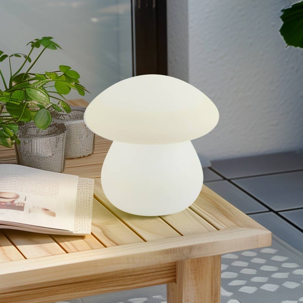 Pilzförmige Tischleuchte auf einem Holztisch im Außenbereich neben einem Buch und einer Pflanze.