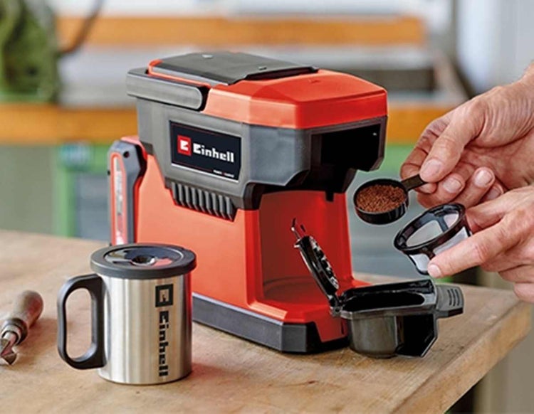 Einhell Akku-Kaffeemaschine auf einem Holztisch mit Edelstahlbecher und Händen, die Kaffeepulver in den Filter füllen.