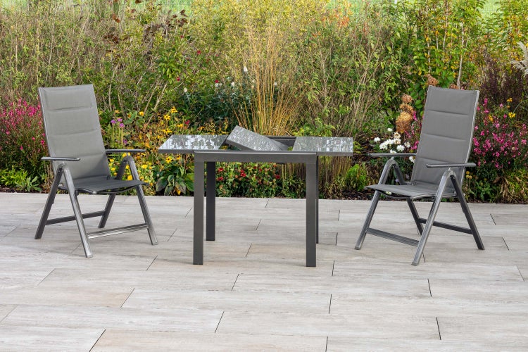 Gartenszene mit zwei Klappsesseln und einem Ausziehtisch mit Glasplatte vor einem bepflanzten Hintergrund.