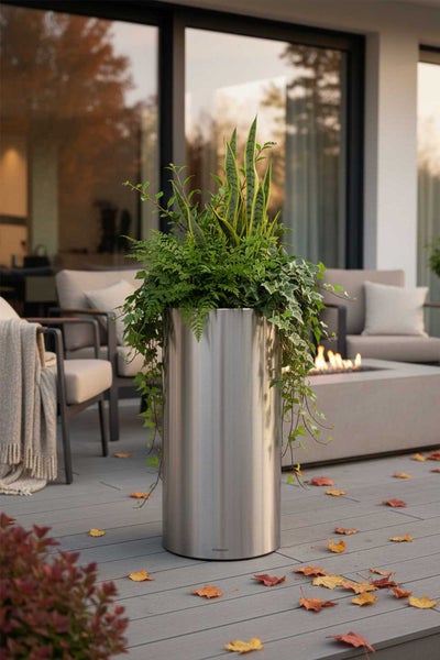 Zylindrischer Pflanzkübel aus Edelstahl mit verschiedenen Pflanzen auf einer hellen Terrasse mit Gartenmöbeln, Feuerstelle und Herbstlaub.