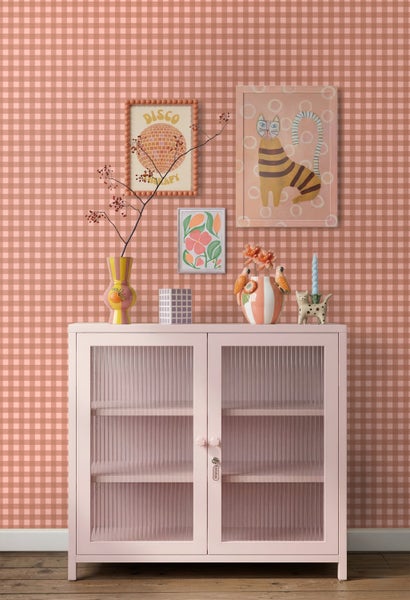 Karierte Tapete in Rosa und Weiß hinter einem rosa Metallschrank mit geriffelten Glastüren und Dekoration in einem hellen, organisierten Raum.