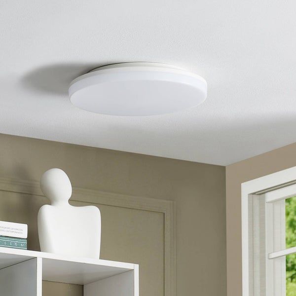 Flache, runde LED-Deckenleuchte in Weiß, montiert an einer weißen Decke über einem Regal in einem hellen, modern eingerichteten Zimmer.