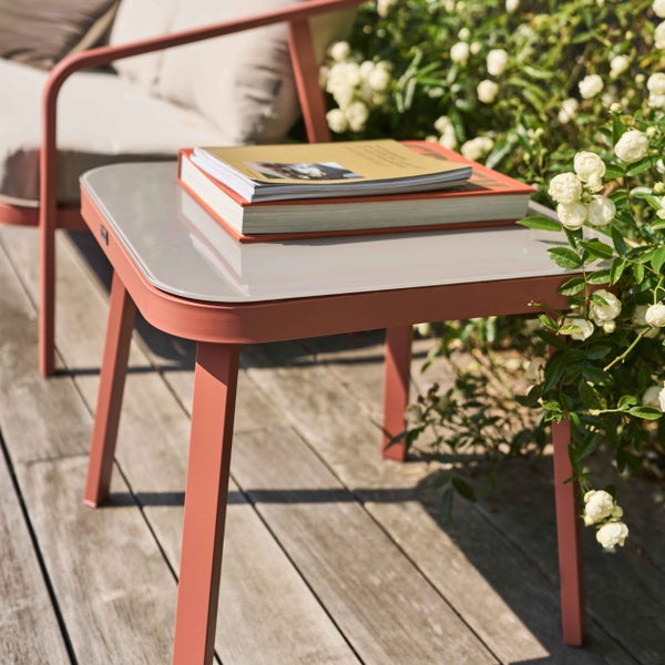 Moderner quadratischer Beistelltisch mit Glasplatte und terrakottafarbenem Metallgestell auf einer Holzterrasse neben einem Rosenstrauch.