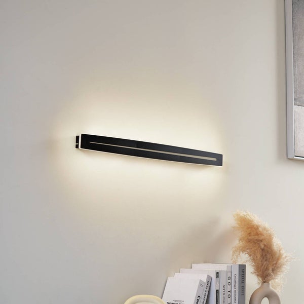 Moderne schwarze LED Wandleuchte mit schmalem Design an einer Wand montiert, eingeschaltet mit warmweißem Licht über Büchern und einer Vase.