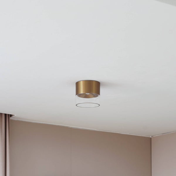 Zylindrischer Deckenstrahler mit messingfarbenem Metallsockel und klarem Glasschirm, montiert an einer Decke.