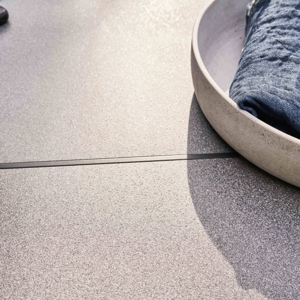 Nahaufnahme einer grauen Terrassenplatte mit strukturierter Oberfläche, einer schmalen Fuge und einer Steinschale mit blauem Tuch.