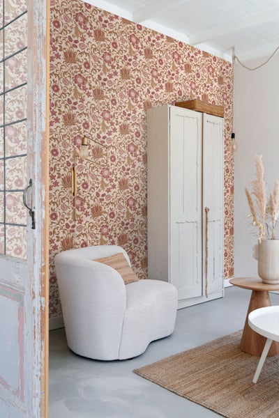 Wohnzimmer mit floraler Tapete, gepolstertem Sessel, Schrank und Holztisch. Heller, organisierter Raum im Vintage-Stil.