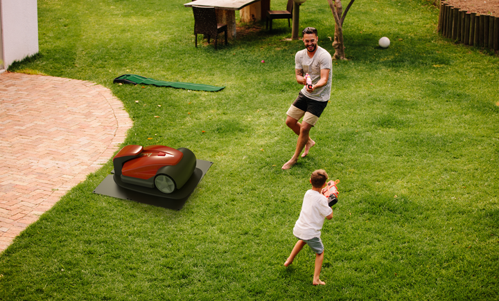 Szene im Garten mit einem Rasenmähroboter, spielenden Kindern und einem Erwachsenen.