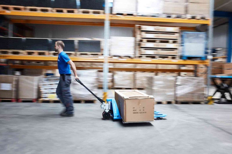 Ein Mitarbeiter zieht einen beladenen Hubwagen durch ein geräumiges Lager mit Hochregalen.