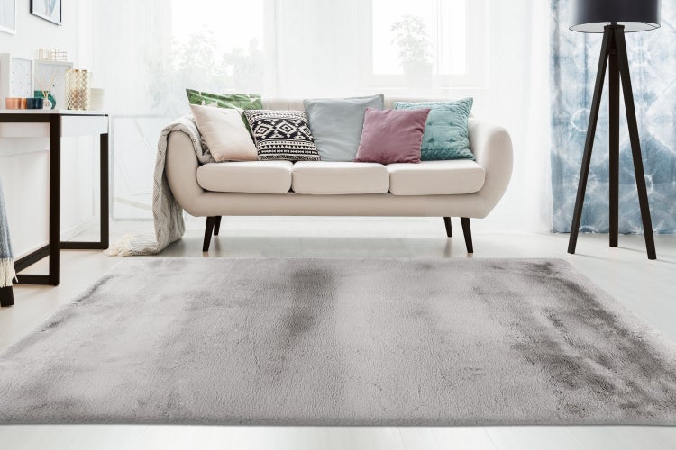 Ein grauer Hochflor Teppich liegt in einem hellen Wohnzimmer vor einem Sofa mit vielen Kissen, einer Stehlampe und einem Konsolentisch.