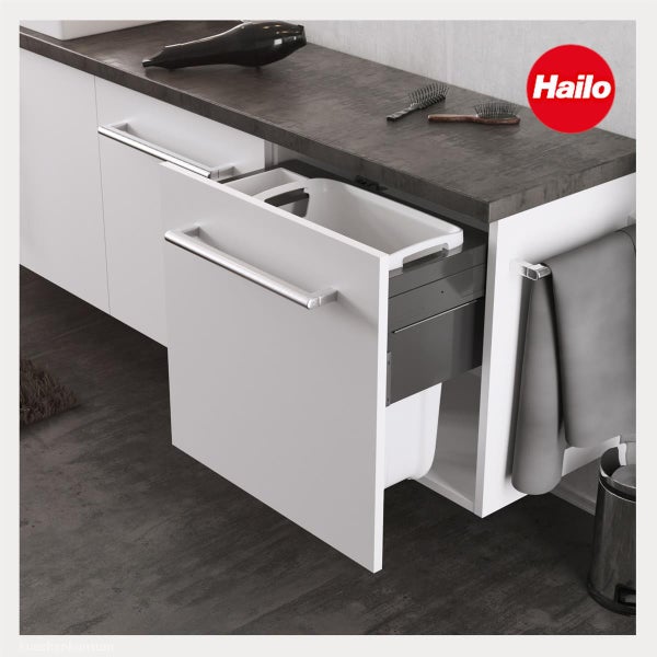 Hailo Logo. Moderner Badezimmerunterschrank mit ausgezogenem Wäschekorb, Haartrockner und Bürsten auf dunkler Arbeitsplatte.