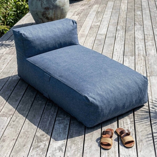 Blaues Outdoor-Liegekissen aus Stoff auf einer hellen Holzterrasse mit Sandalen und einem Pflanzgefäß im Hintergrund.