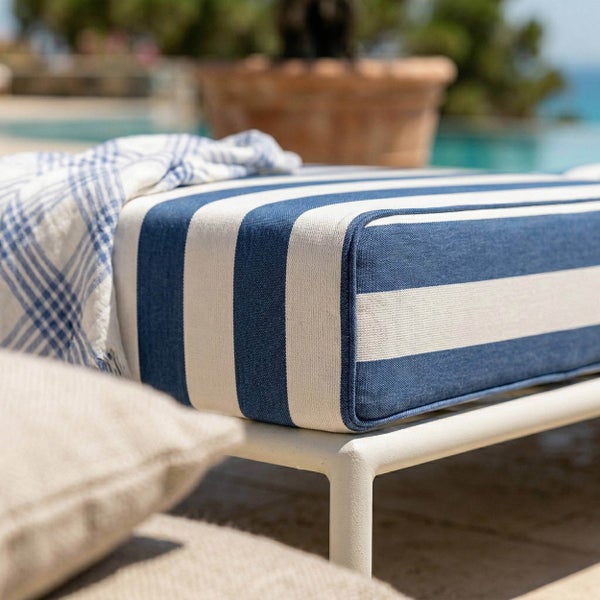 Blau-weiß gestreiftes Loungekissen für den Außenbereich auf einem weißen Metallrahmen in einem hellen Poolbereich.