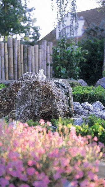 Naturstein-Quellstein in einem Garten mit sprudelndem Wasser, umgeben von Kieselsteinen und Pflanzen vor einem Bambuszaun.