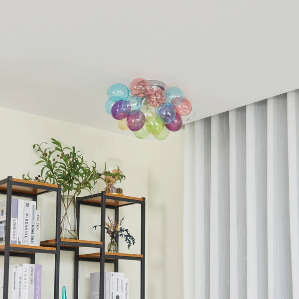 Moderne Deckenleuchte mit bunten Glaskugeln in einem hellen Raum über einem schwarzen Metallregal mit Büchern und einer Pflanze.