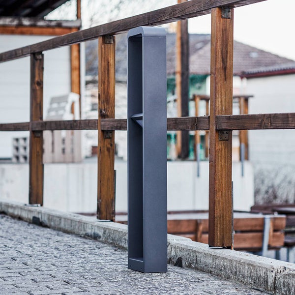 Moderne, rechteckige Wegeleuchte aus anthrazitfarbenem Metall im Außenbereich neben einem Holzgeländer auf einem gepflasterten Boden.