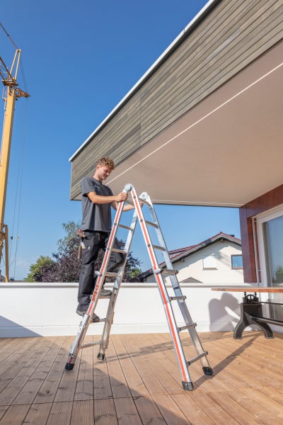 Ein Mann steht auf einer Mehrzweckleiter vor einem modernen Gebäude unter blauem Himmel.