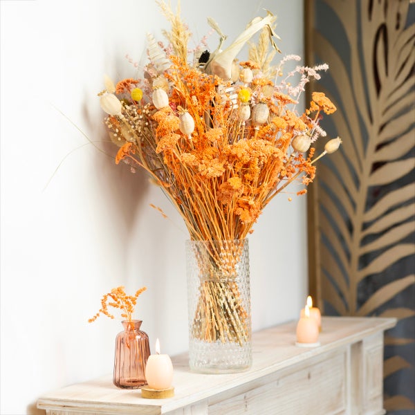 Großer Strauß aus Trockenblumen in einer strukturierten Glasvase auf einem Sideboard aus Holz mit brennenden Kerzen.