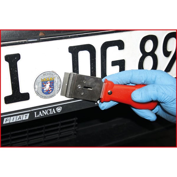 Eine Hand mit Handschuh entfernt mit einem Schaber Aufkleber vom Nummernschild.