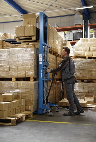 Ein Mann bedient einen blauen Hochhubwagen in einem Lagerhaus, um Kartons auf einer Palette anzuheben.
