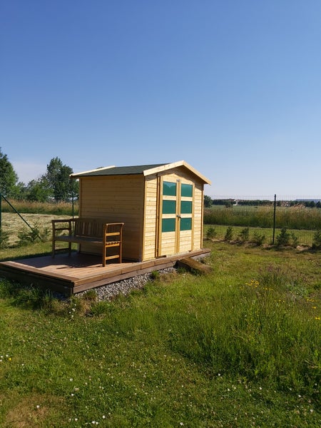 Gartenhaus aus hellem Holz mit Doppeltür, vier Fensterscheiben pro Türflügel und kleiner Holzterrasse mit Bank auf einer Rasenfläche.