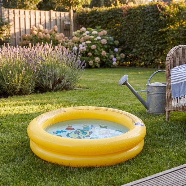 Aufblasbares Planschbecken für Kinder mit Fischmotiven auf dem Rasen im Garten neben einer Gießkanne und einem Korbsessel.