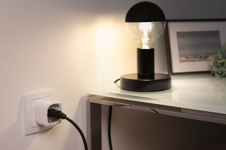 Smarter Zwischenstecker in einer Wandsteckdose, verbunden mit einer modernen Tischlampe auf einem Glastisch in einer hell beleuchteten Umgebung.