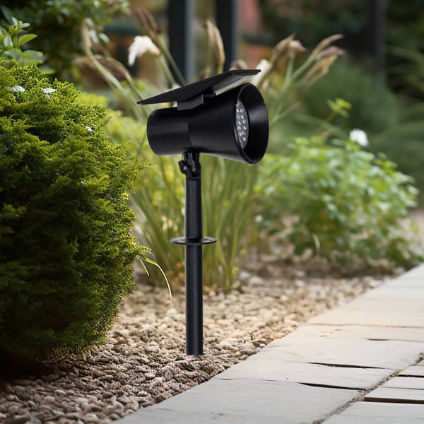 Schwarzer LED-Solarstrahler mit Erdspieß und Solarpanel, installiert in einem Garten neben einem gepflasterten Weg.